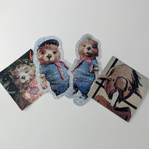 May include: Three glitter stickers featuring characters from the film Paddington. The stickers are on a white background. The first sticker shows Paddington wearing a blue and white sailor outfit. The second sticker shows Paddington wearing a blue and white sailor outfit. The third sticker shows a brown bear with a toothy grin.
