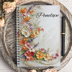 Pode incluir: Um caderno de espiral com fundo branco e um design floral em tons de rosa, laranja, amarelo e roxo. O texto "Penelope" está escrito em cursiva preta na capa.