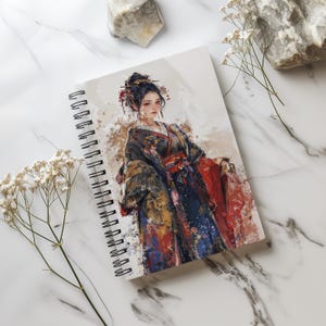 Può includere: Un quaderno a spirale con un'illustrazione ad acquerello di una donna in un kimono colorato. La donna ha i capelli scuri e indossa un kimono rosso e oro con un motivo floreale. Lo sfondo è un acquerello blu chiaro.
