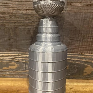 Puede incluir: Un trofeo plateado con una parte superior detallada en forma de cuenco acanalado y una base escalonada. El trofeo es cilíndrico con bandas horizontales y una superficie texturizada. El fondo es una superficie de madera.