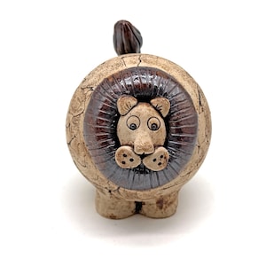 Può includere: Statuetta di leone in ceramica con corpo rotondo e criniera marrone scuro. Il leone ha un viso marrone chiaro con grandi occhi e un naso piccolo. Anche la coda è marrone scuro. La statuetta ha un aspetto strutturato e screpolato.