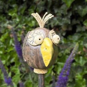 Könnte beinhalten: Ein runder Keramik-Gartenstecker in Vogelform mit dunkelbrauner Metallic-Glasur, einem leuchtend gelben Schnabel und zwei großen, hervorstehenden Augen mit weißem Rissmuster. Der Vogel hat ein kleines Büschel heller Fasern auf dem Kopf.