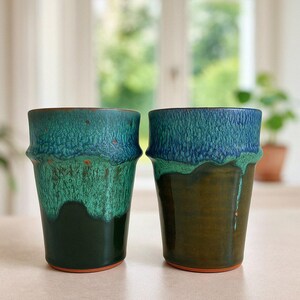 Peut inclure: Deux mugs en céramique avec une finition émaillée texturée. Les mugs présentent un dégradé de couleurs, passant du vert foncé au bleu sarcelle et au bleu. Ils ont une forme cylindrique avec un bord légèrement évasé, parfaits pour les boissons chaudes.