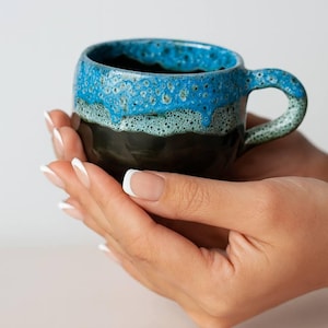 Puede incluir: Una taza de cerámica hecha a mano con un diseño único. La taza presenta una base oscura, una sección media verde y una sección superior azul texturizada. El asa es de color azul-verdoso a juego.