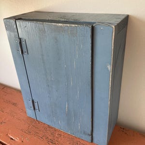 May include: A small, blue wooden cabinet with a single door and two hinges. The cabinet has a distressed, rustic look and is made of reclaimed wood.