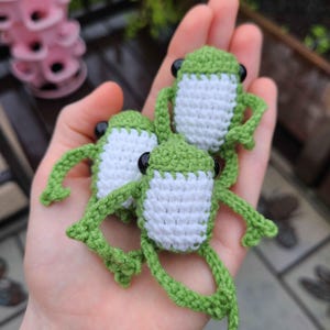 Puede incluir: Tres ranas de crochet verdes con vientres blancos y ojos negros. Las ranas están sentadas en la mano de una persona.