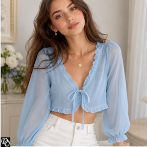 May include: A light blue sheer cropped blouse with long puff sleeves, a deep V-neckline, and ruffled trim along the edges. The front features a delicate tie closure at the center, and the fabric has a soft, translucent quality throughout the garment.