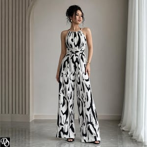 Könnte beinhalten: Ein ärmelloser Jumpsuit mit hohem Neckholder-Ausschnitt und weit geschnittenen Hosenbeinen. Das Kleidungsstück besteht aus weißem Stoff mit einem markanten, abstrakten schwarzen Pinselstrichmuster. Es hat eine betonte Taille und ein Bindedetail.