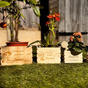 May include: Four wooden planter boxes with personalised messages. The boxes are decorated with a cartoon image of a person holding a plant. The messages read "From your grandson James", "From your daughter Charlotte", "Andrea", and "From your son James".