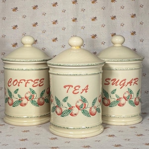 May include: Three cream ceramic storage jars with lids, labelled "COFFEE", "TEA", and "SUGAR" in red script. Each jar has a decorative band of red apples and green leaves. The lids have a small knob on top.