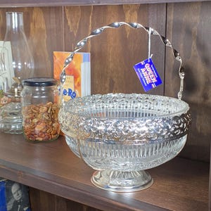May include: A clear glass serving bowl with a silver rim and handle. The bowl has a pedestal base and a decorative pattern around the rim. A blue tag hangs from the handle. The bowl is on a wooden shelf.
