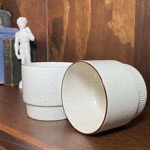 May include: Two speckled white ceramic mugs with a brown rim and a ribbed base. One mug is upright, the other is on its side. The mugs are on a wooden surface.