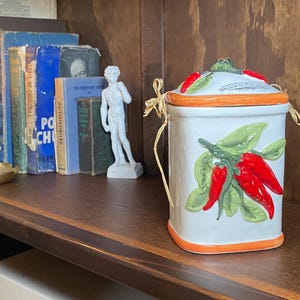 May include: A white ceramic jar with a lid, decorated with red chilli peppers and green leaves. The jar has an orange rim and is tied with raffia. Books, a candle, and a small statue are in the background.