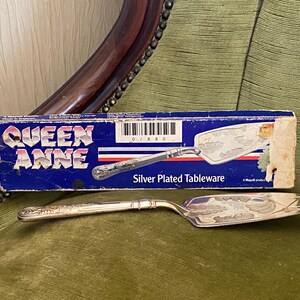 May include: A silver-plated cake server with an ornate handle and a decorative blade, next to its original blue box. The box reads "QUEEN ANNE" and "Silver Plated Tableware". The server is displayed on a green upholstered surface.