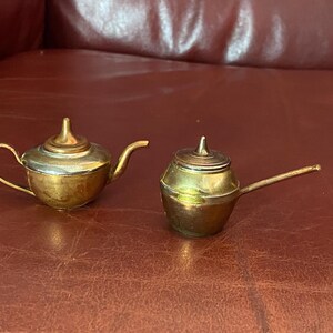 May include: Two small, antique brass teapots. One has a curved handle and a silver-coloured top. The other has a long, straight spout and a lid. Both are on a brown surface.