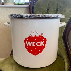 May include: A large white enamel preserving pot with two side handles and a clear plastic lid. A red strawberry logo with the word "WECK" is on the front. The pot is on a green upholstered chair.