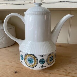 May include: White ceramic coffee pot with a lid, handle, and spout. The base has a repeating pattern of teal, yellow, and black circular designs. A matching white ceramic container is in the background.