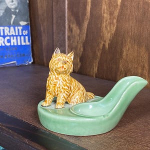 May include: A ceramic figurine of a golden-brown terrier dog sitting on a light green, oval-shaped base that features a smooth, curved indentation designed to hold a small object, with a glossy finish on both the dog and the base.
