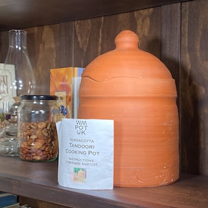 May include: A terracotta tandoori cooking pot with a lid, on a wooden shelf. A jar of seeds and a glass bottle are also on the shelf. A paper with instructions is next to the pot.