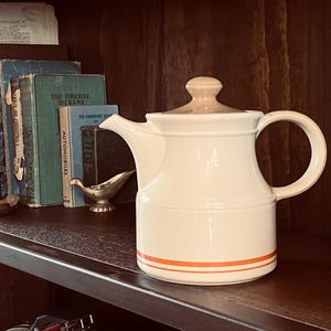 May include: A white ceramic teapot with a tan lid and handle, featuring a thin orange stripe. The teapot sits on a dark wooden shelf with vintage books, a candle, and a small metal dish. The scene has a classic, charming aesthetic.