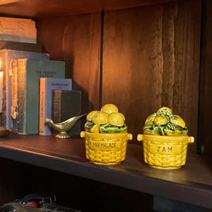 May include: Two yellow ceramic jars, labelled "Marmalade" and "Jam", sit on a shelf. The jars are basket-shaped and topped with fruit. A lit candle, books, and a small brass bird figurine are also on the shelf.