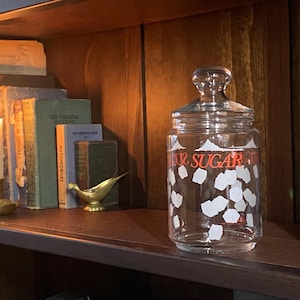 May include: A clear glass jar with a lid, labelled "SUGAR" in red lettering, and decorated with white shapes. The jar sits on a dark shelf with books, a candle, and a brass bird figurine. The background is a wooden cabinet.
