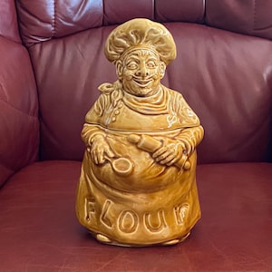 May include: A tan ceramic flour container shaped like a smiling chef wearing a toque and apron. The apron has the word "FLOUR" on it. The chef holds a rolling pin and a spoon. The container is on a red surface.