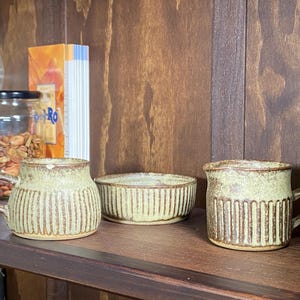 May include: Three ceramic items: two mugs and a small bowl. The mugs and bowl are light green with vertical brown stripes. The mugs have handles. The items are on a dark brown wooden shelf.