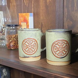 May include: Two ceramic mugs with a light green glaze and a brown, circular design. The mugs have a handle and a beige base. They are displayed on a wooden shelf with other items, including a glass jar and a book.
