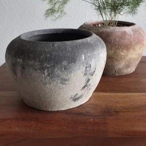 May include: Two weathered, round ceramic planters. One is dark gray with a lighter, textured base. The other is a reddish-brown with a plant featuring delicate, green foliage. Both sit on a wooden surface, suggesting a rustic, natural aesthetic.