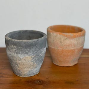 May include: Two small, weathered terracotta pots. One is painted gray with a textured, aged appearance. The other is a natural orange-brown color, also with a textured surface. Both are empty and sit on a wooden surface.