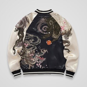 Puede incluir: Una chaqueta bomber de satén negra y color crema con bordados intrincados de dos dragones, nubes arremolinadas y pequeñas flores de cerezo rosas. La chaqueta tiene mangas largas color crema, cuello, puños y dobladillo acanalados en negro.