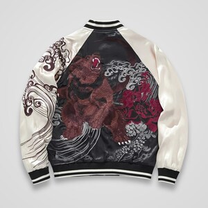 Puede incluir: Una chaqueta bomber de satén negra y crema con un gran bordado detallado de un oso pardo rugiendo en la espalda. El diseño incluye intrincados patrones de olas blancas en las mangas y un grupo de elementos florales de color rojo oscuro junto al oso.