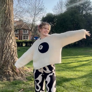 Puede incluir: Una persona con un suéter color crema con un gran gráfico de bola ocho negra. La persona también usa pantalones estampados en blanco y negro y zapatillas blancas. El fondo es una escena al aire libre soleada con un árbol y hierba verde.