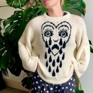 Puede incluir: Suéter color crema con un gráfico en forma de corazón negro con ojos, bigote y lágrimas. El suéter tiene mangas largas y cuello redondo. La persona lleva pantalones de lunares blancos y negros.