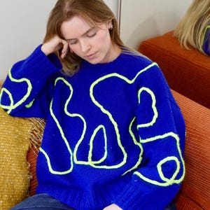 以下が含まれることがあります： ネオングリーンの抽象的なデザインが施された青いセーター。セーターはクルーネックで、ニット素材です。その人は青いジーンズを履いています。