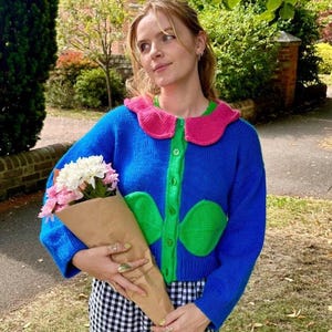 Peut inclure: Un cardigan bleu avec un col rose et des détails verts. Le cardigan a des poches vertes en forme de fleur et des boutons verts. La personne tient un bouquet de fleurs dans du papier brun. Elle porte un haut vert et une jupe à carreaux noirs et blancs.