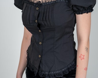 Puede incluir: Una blusa negra de manga corta con ribete de encaje y cierre de botones. La blusa tiene un corpiño ajustado y una manga ligeramente abullonada. La modelo lleva una falda negra con ribete de encaje.