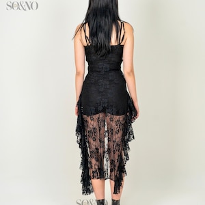 May include: A black lace dress with a ruffled hemline. The dress is sheer and has a fitted bodice. The model is wearing black heeled boots.