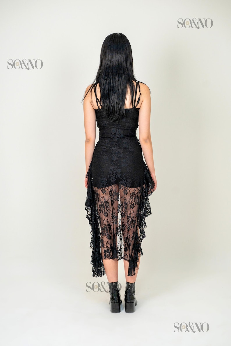 May include: A black lace dress with a ruffled hemline. The dress is sheer and has a fitted bodice. The model is wearing black heeled boots.