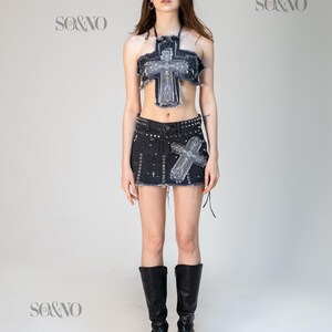May include: A denim two-piece set featuring a cross design. The set includes a halter top and a mini skirt, both with distressed edges and embellished with a cross. The model is wearing black knee-high boots.