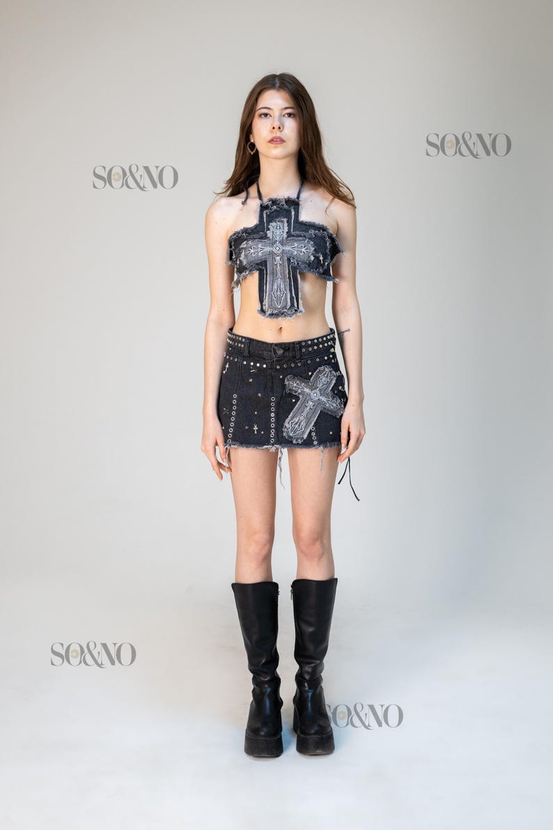 May include: A denim two-piece set featuring a cross design. The set includes a halter top and a mini skirt, both with distressed edges and embellished with a cross. The model is wearing black knee-high boots.