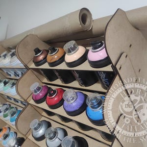 Puede incluir: Un organizador de pintura en aerosol de madera con m&uacute;ltiples compartimentos, que contiene varias latas de pintura en aerosol de colores. Las latas tienen bases negras y tapas de colores. Un rollo de papel est&aacute; montado encima del organizador.