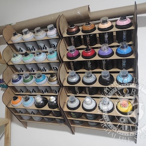 Puede incluir: Estante de almacenamiento de latas de pintura en aerosol de madera lleno de varias latas de pintura en aerosol. Las latas son de diferentes colores, incluyendo rosa, azul y dorado. El estante es de madera marrón claro y está montado en una pared blanca.