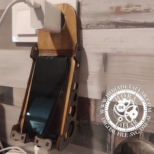 May include: Wooden phone charging station with a dark phone in place. The station is mounted on a wall and has a cutout design. A white charger is plugged into the wall and the phone. The station has the text "HORA DETALLAR" and "DIGITAL FILE SVG-DXF-AI".