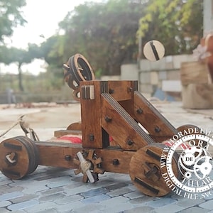 May include: A wooden toy catapult with a string attached to the arm. The catapult is made of brown wood and has a black and white wheel. The catapult is on a grey and white tiled surface. The text "www.horadetalla.com DIGITAL FILE SVG DOWNLOAD" is visible on the surface.