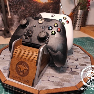 May include: A wooden stand with a gray and black camouflage Xbox controller and a white Xbox controller. The stand has a logo that says "Dauntless" and a circular design with the text "Hora de Tallar" and the text "@horaddetallar".