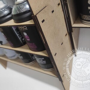 Puede incluir: Un organizador de latas de pintura en aerosol de madera con m&uacute;ltiples estantes. El organizador contiene varias latas de pintura en aerosol, incluidas marcas como PRO y MARLACK. Las latas son negras, blancas y plateadas. El organizador es de madera marr&oacute;n claro.