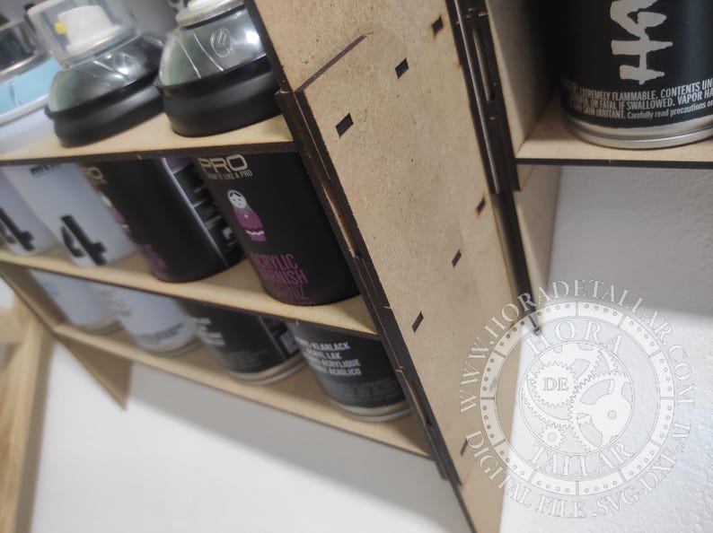 Puede incluir: Un organizador de latas de pintura en aerosol de madera con m&uacute;ltiples estantes. El organizador contiene varias latas de pintura en aerosol, incluidas marcas como PRO y MARLACK. Las latas son negras, blancas y plateadas. El organizador es de madera marr&oacute;n claro.