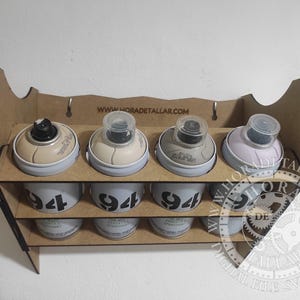 Puede incluir: Un organizador de madera para botes de pintura en aerosol con cuatro botes en cada uno de los dos niveles. Los botes son de color beige, gris y lila claro. La fila inferior de botes tiene el n&uacute;mero "94" impreso. El sitio web "www.horadetallar.com" est&aacute; impreso en la parte superior.